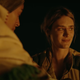 A woman and a tall man, both in jackets, warm themselves by a campfire.